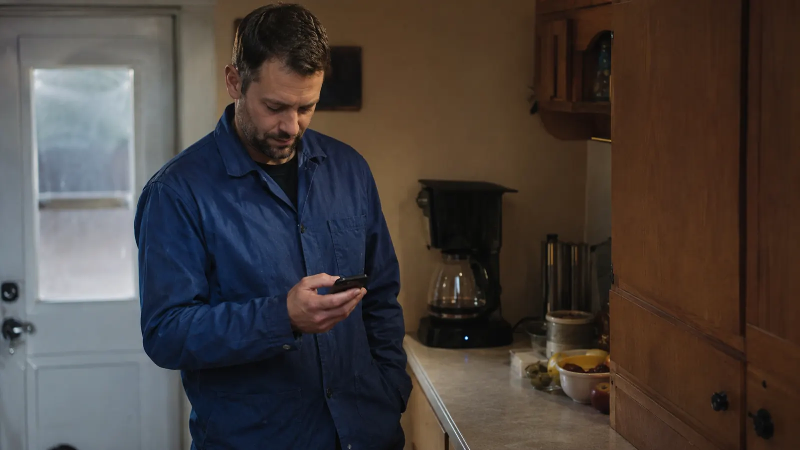 A quiet galley kitchen at early morning with a husband in his coat checking his phone while a full pot of coffee brews