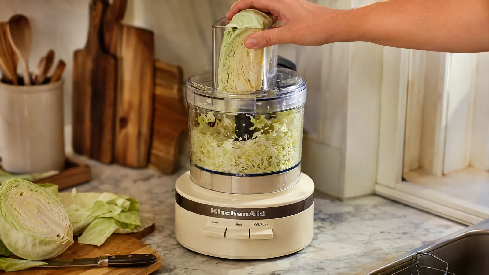 A hand feeding a head of cabbage into a food processor's feed tube on a narrow kitchen counter